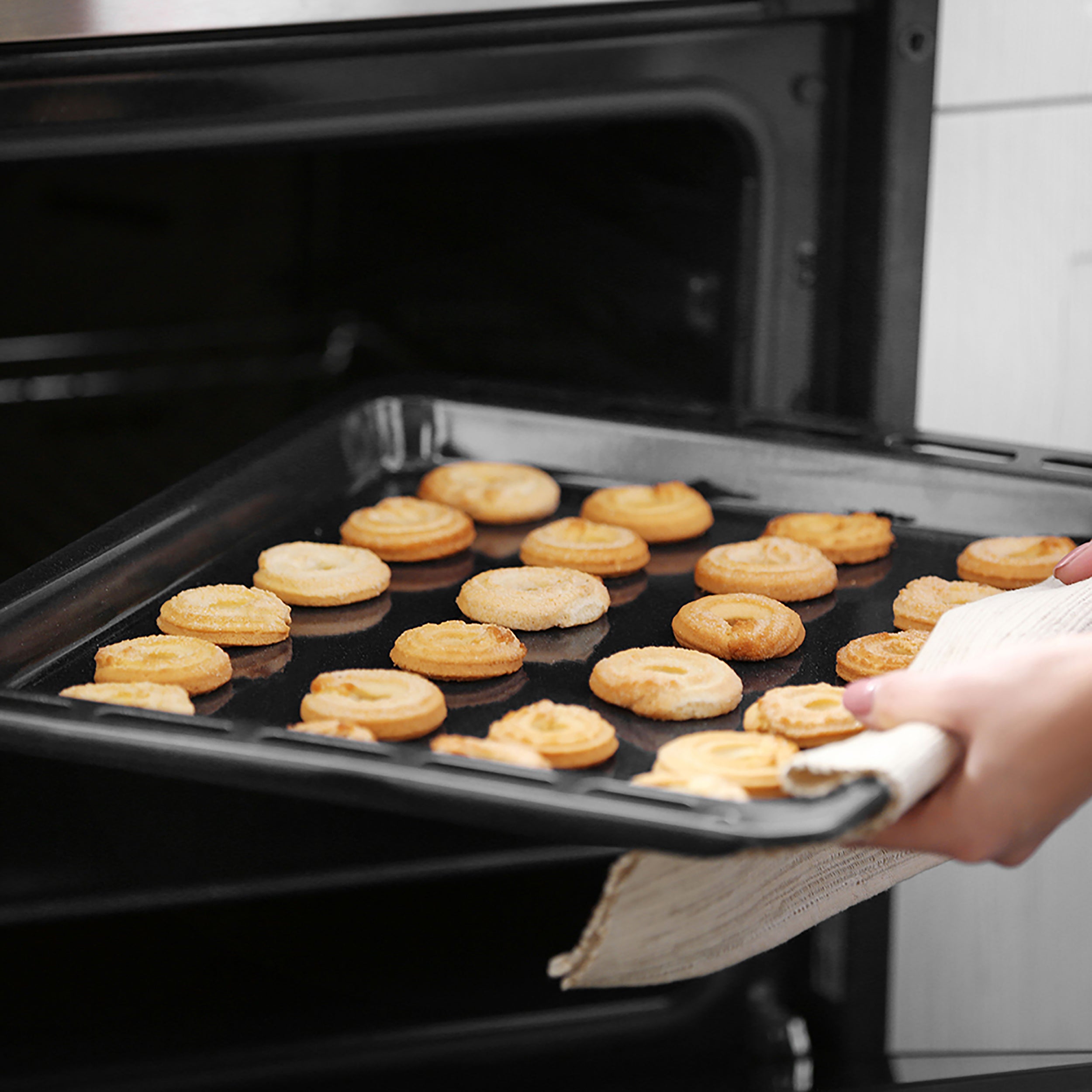 A person removes an Oven Baking Tray compatible with the Empava 24-inch Single Wall Oven, filled with small, golden-brown, swirl-shaped pastries from an oven. The tray is being held with a white oven mitt, and the freshly baked pastries appear appetizing.