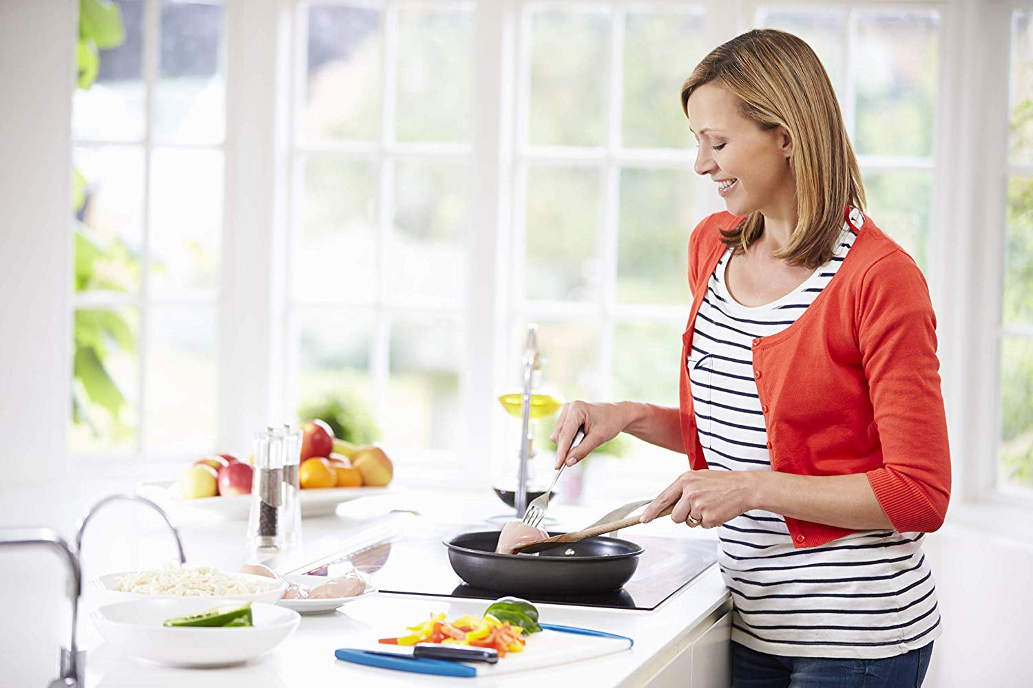 A woman wearing a red cardigan and striped shirt smiles while cooking in a bright kitchen adorned with Empava appliances. She is using a spatula to flip food in a frying pan on the Empava 12 In. Induction Cooktop with 2 burners. In the foreground, various chopped vegetables and kitchen utensils are visible on the counter.