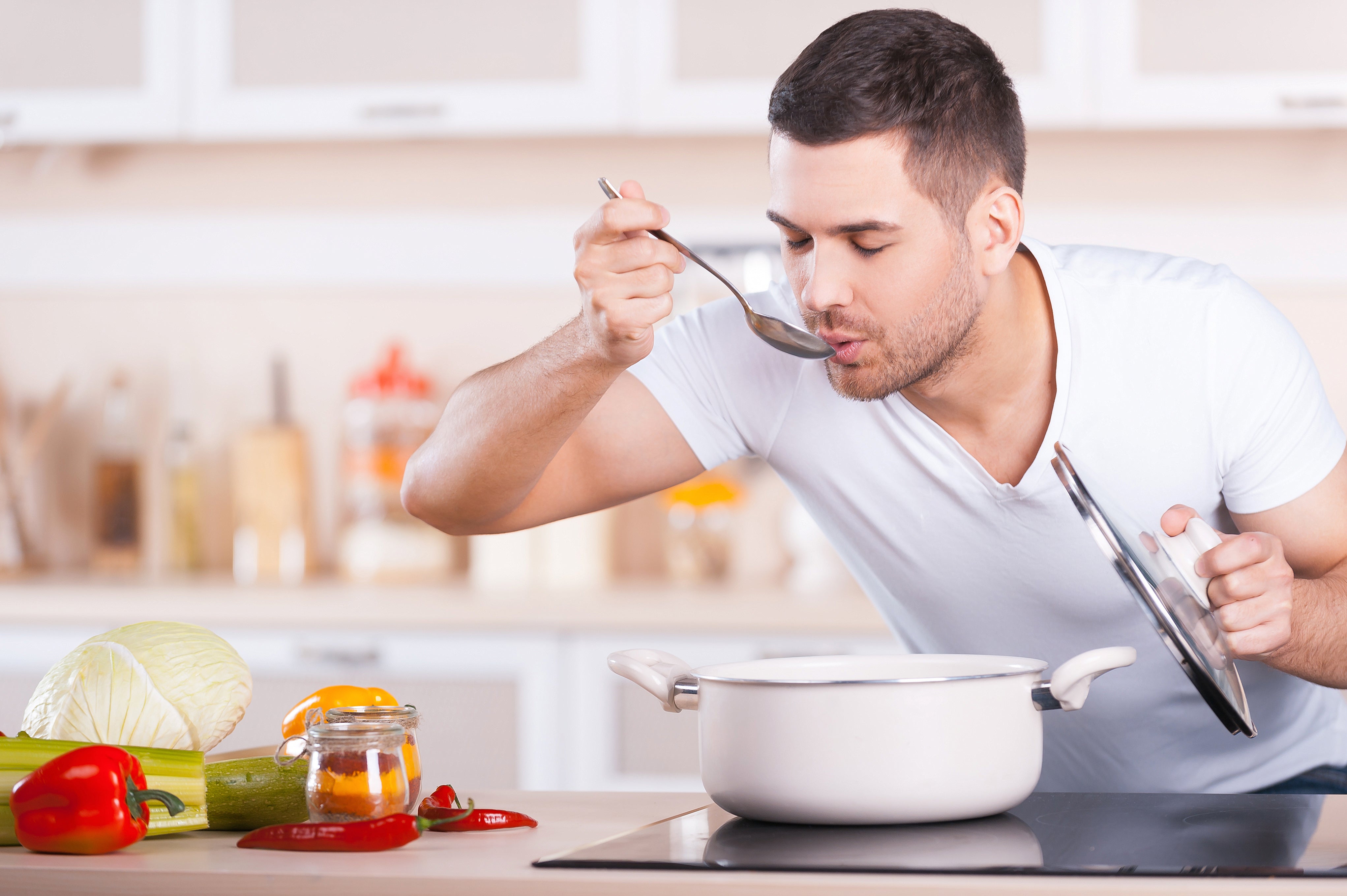 A man with short dark hair in a white t-shirt is tasting soup from a spoon in a modern kitchen equipped with an Empava 24 in. W x 20.5 in. D Induction Cooktop. He is holding the pot lid in one hand and blowing on the spoon, with fresh vegetables, including cabbage and bell pepper, on the counter beside him.