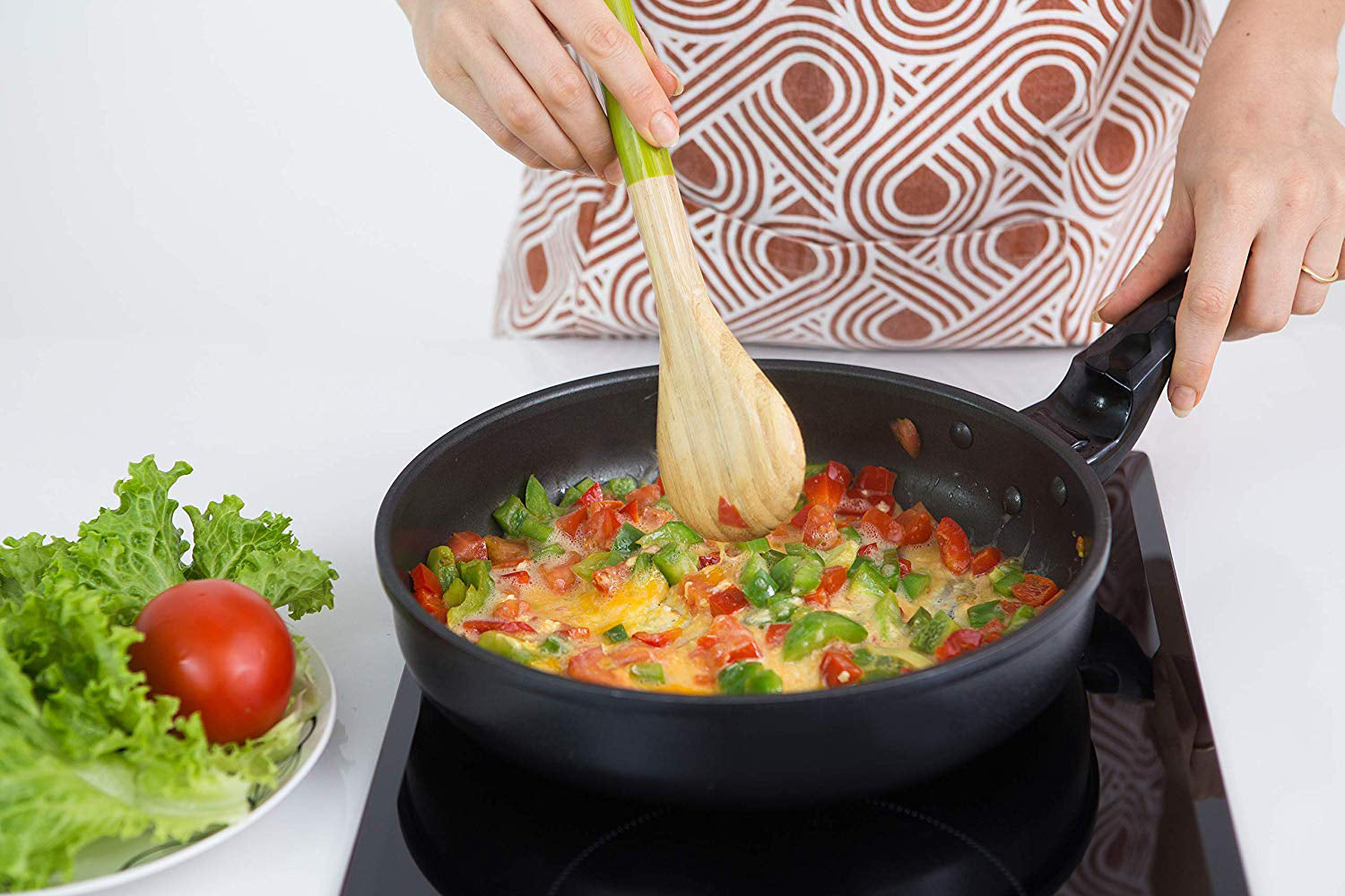 A person cooking an omelette in a black frying pan on an Empava 12 inch Portable Induction Cooktop, using a wooden spoon with chopped tomatoes and green bell peppers. In the foreground, there's a plate with fresh lettuce and a whole tomato on a white surface. The person, wearing a patterned apron, is standing next to the sleek Empava appliance for added convenience.