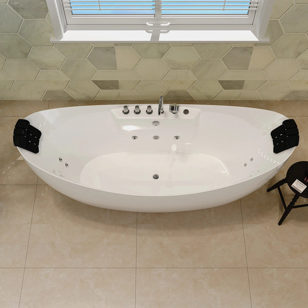 A modern bathroom featuring beige-tiled floors and hexagonal wall tiles is graced by the 67" Freestanding Boat-shaped Whirlpool Bathtub with a Center Drain, characterized by its white color and oval shape. The tub includes black headrests on both sides for added comfort. Silver fixtures are installed on one side of the crack-resistant acrylic tub, and a small stool with a book sits nearby, completing the serene setting.