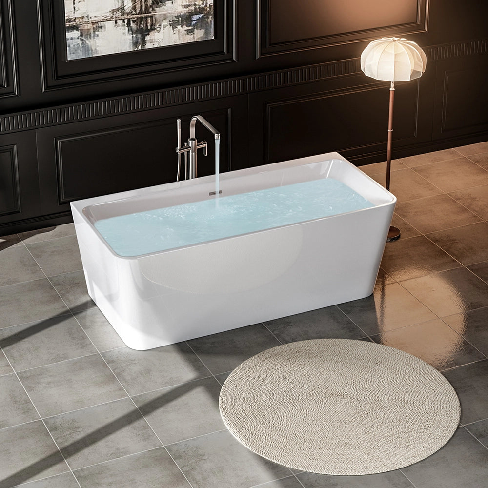 A modern bathroom featuring the 67" Freestanding Soaking Tub with Center Drain, filled with water. The tub's ergonomic design and durable acrylic material ensure comfort and longevity. The floor is tiled in light gray, with a round beige rug in front of the tub. A tall lamp illuminates the space, and an abstract painting hangs on the dark-paneled wall.