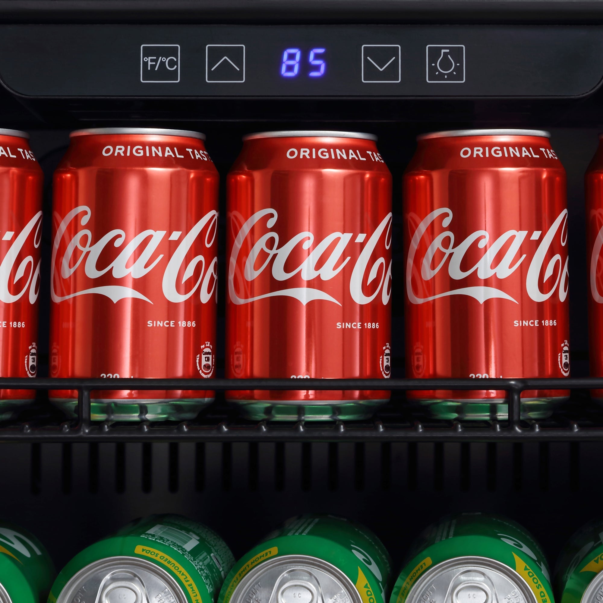 An Empava 24 Inch Freestanding & Under Counter Beverage Fridge shelf holds red cans of Coca-Cola neatly arranged in a row. Below them, vibrant green and yellow cans add a pop of color. The refrigerator’s digital thermometer displays "85" in blue.