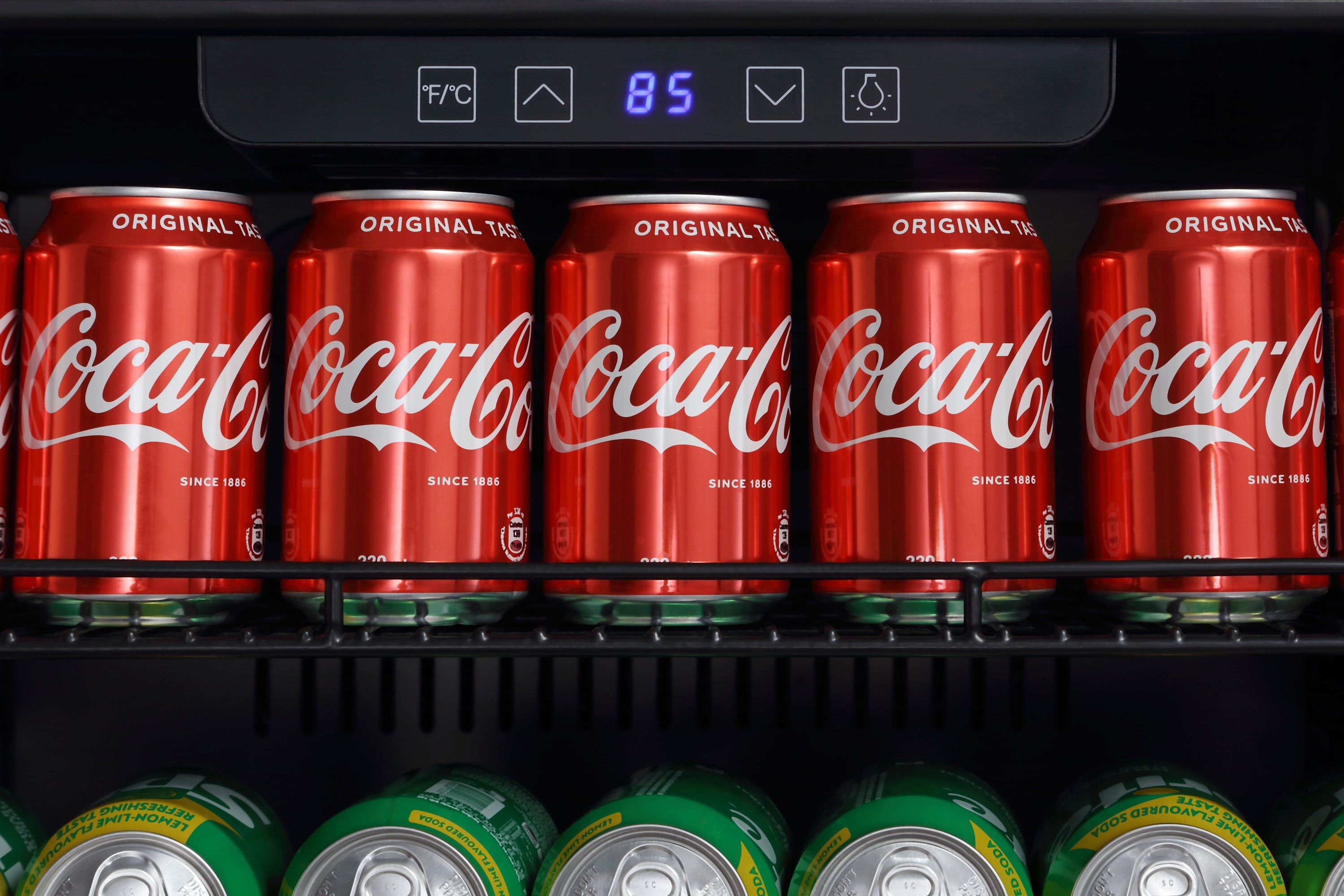 An Empava 24 Inch Freestanding & Under Counter Beverage Fridge is displayed, filled with soda cans to illustrate the high quality of Empava appliances. The top shelf features red Coca-Cola cans neatly arranged, with an electronic display reading "8.5" above them. The bottom shelf showcases a row of green cans partially visible from below.