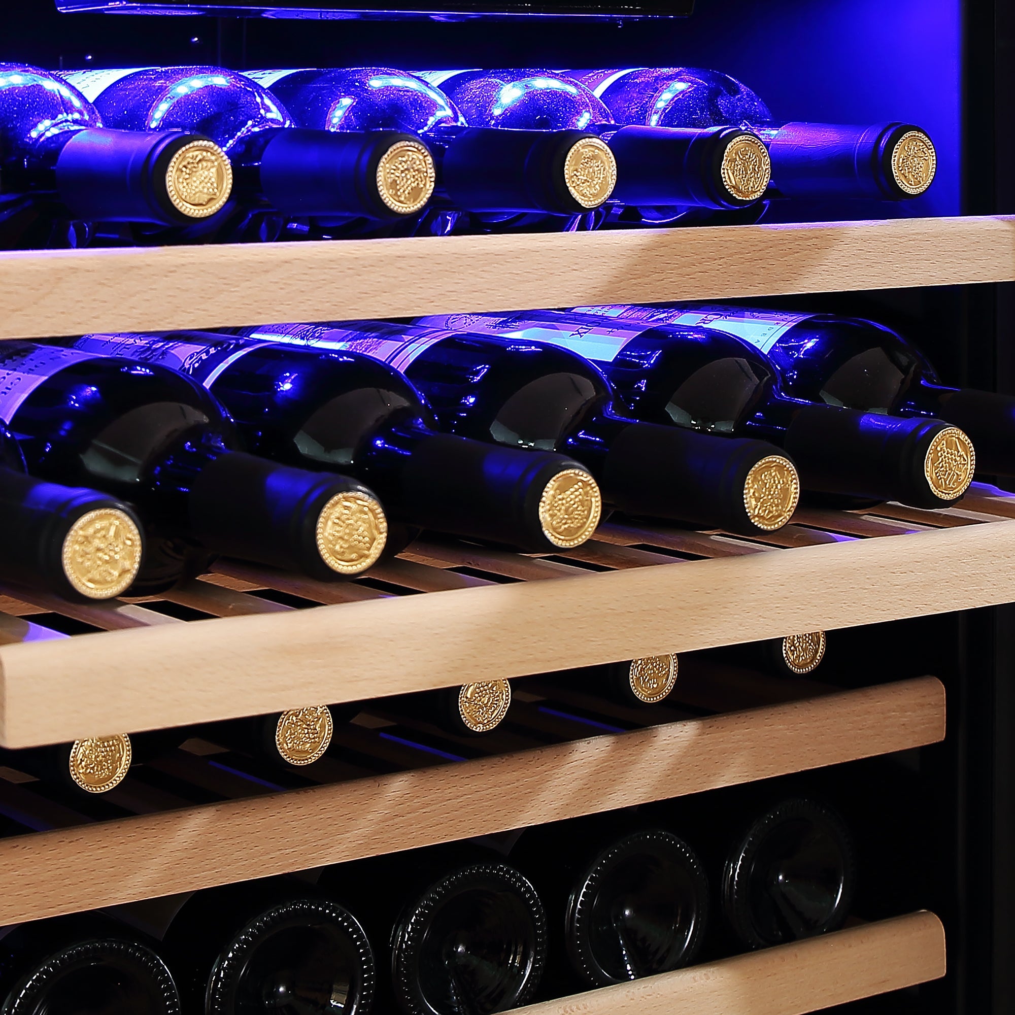 Close-up of an Empava 24" Wine Cooler 55" Tall Wine Refrigerator filled with rows of wine bottles laying horizontally on wooden shelves. The bottles are arranged with their necks facing outwards, and are illuminated by a blue light from above, highlighting the golden seals on the bottle tops.