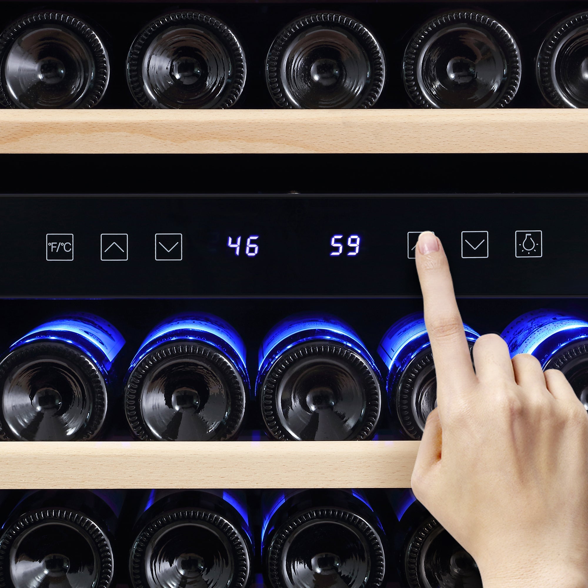 A person adjusts the settings on the dual zone digital control panel of an Empava 55" Tall Dual Zone 100 Bottle Wine Fridge filled with wine bottles. Two temperature settings are visible on the display: 46°F and 59°F. The fridge features wooden shelves and blue interior lighting that highlights the bottles, helping to prevent wine oxidization.