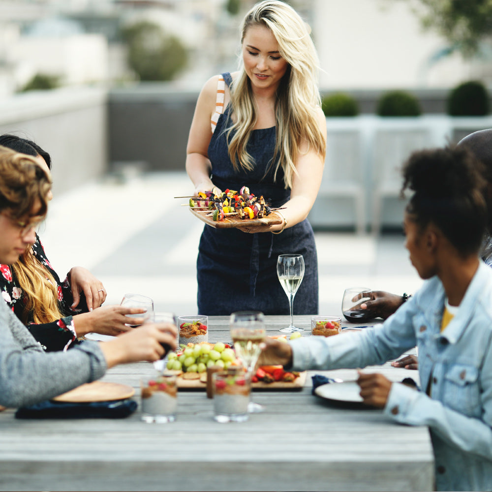 A woman in a denim apron serves kabobs to a group of people seated at an outdoor dining table, which is equipped with the Empava 5 Feet Propane Adapter Hose 1 lb to 20 lb with Upgraded Gauge EMPV-50EH47. The table is set with various dishes, glasses of wine, and a large bowl of grapes. The atmosphere appears casual and friendly, with everyone engaged in conversation.