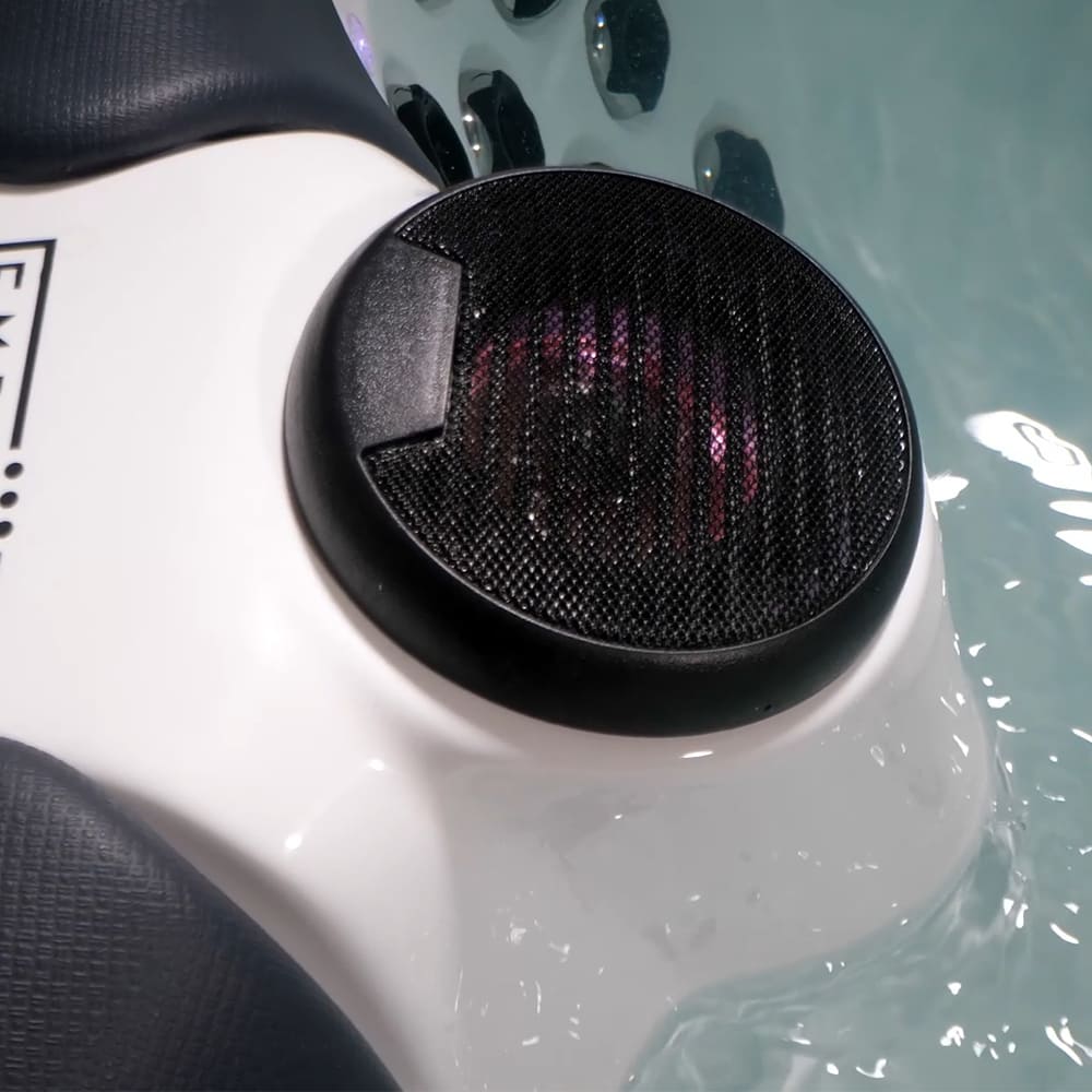 A close-up image of an Empava 75 in. Freestanding Luxury Thermostatic 2-Person Rectangle LED Tub with Right Drain shows a circular, black mesh-covered waterproof speaker mounted on a pet-friendly flotation device in the tub. Faintly glowing purple LED lights shine underneath the speaker's mesh cover, while the water, benefiting from ozone disinfection, gently ripples around the non-toxic device.