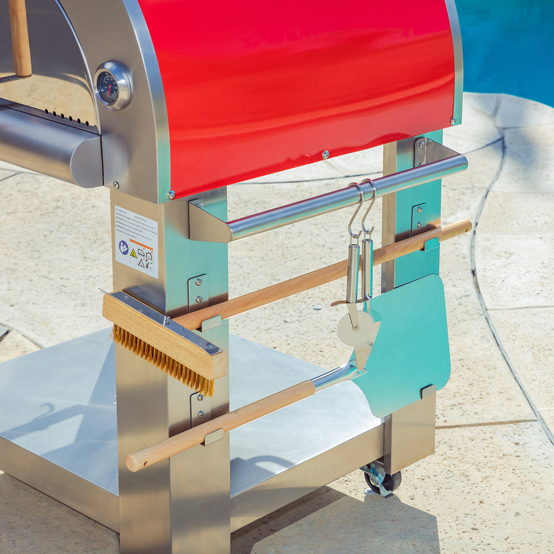 An **Empava Outdoor Wood Fired Pizza Oven** with a vibrant red top, part of an All-in-One Oven Kit, situated on a patio by the pool. Attached are various wooden-handled tools, including a brush, pizza peel, and spatula, all neatly hanging on metal brackets to achieve the perfect wood-smoked flavor.