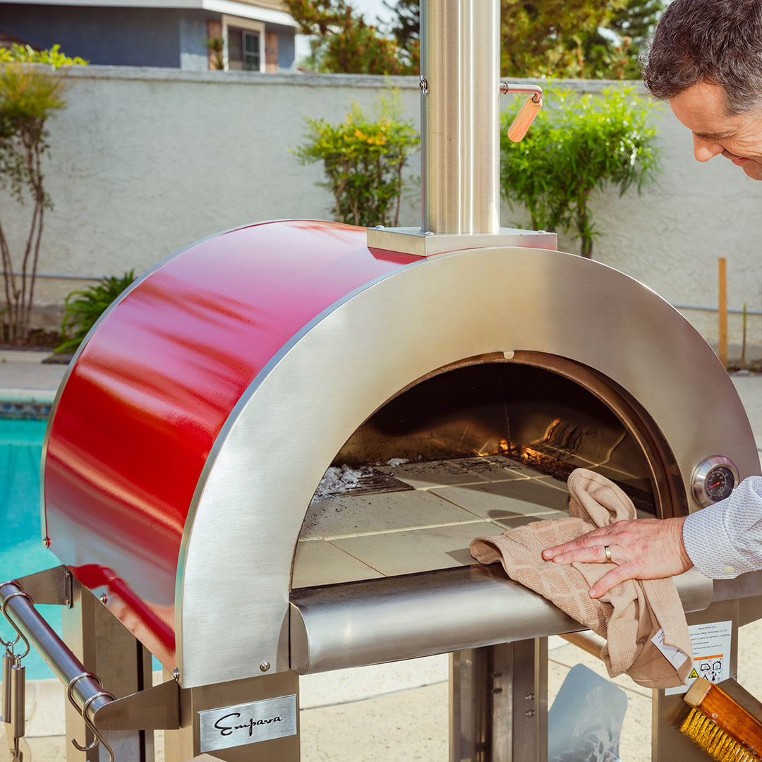 A person cleans the interior of an Empava Outdoor Wood Fired Pizza Oven, which features a red top. The oven, ideal for achieving that authentic wood-smoked flavor, is positioned by a swimming pool in a backyard. The person uses a cloth and is focused on the task.