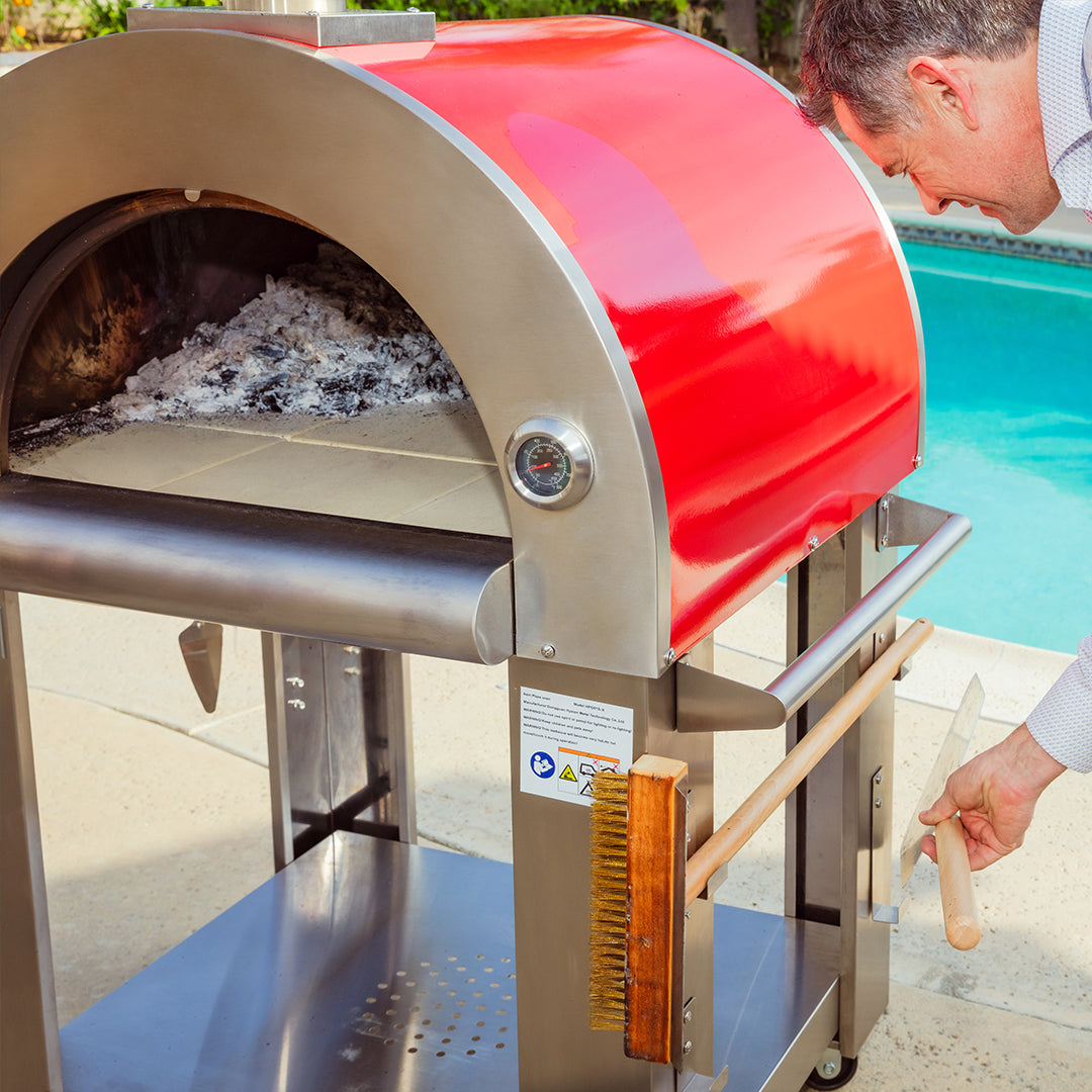 A person uses a wooden brush to clean the Empava Outdoor Wood Fired Pizza Oven, which features a stainless steel finish and a red dome, situated near a swimming pool. Ashes inside suggest recent use, enhancing the wood-smoked flavor. The pizza oven includes a built-in thermometer and is set up on a paved patio area, creating an all-in-one oven kit dream setup.